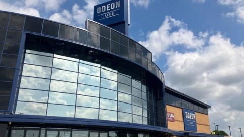 The outside of the cinema. It has curved reflective glass on the front and a sign above it saying Odeon Luxe