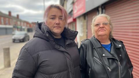 Two women stand together on a quiet high street. One has pink hair and a padded black coat. The other older woman wears glasses and a leather jacket