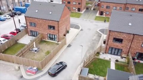 an ariel view of a housing estate with unfinished roads, rubble and a black car driving into the estate.