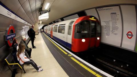 A platform at Knightsbridge Tube station, with a Tube train pulling up to the platform. A few people are waiting on the platform, some sitting on a bench.