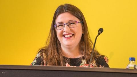 A woman with long brown hair wears purple-framed glasses. She has a black top with floral pattern. She is smiling against a yellow backdrop, stood in front of a microphone.