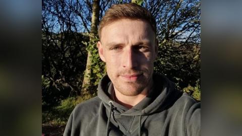 David Law looking straight at the camera. He is wearing a grey hoodie. He is stadning in front of a bare tree and some greenery. He has short, fair, brown hair and short stubble.