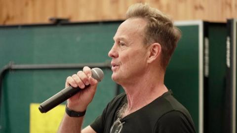 Jason Donovan, wearing a black T-shirt, holding a microphone and singing into it.