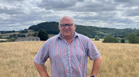 Martin Williams stood in his field which is filled with brown, dry crops. He is wearing a red, blue and white checked short-sleeve shirt and is also wearing black square glasses and has a bald head. 