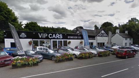 A flat roofed car show room on the side of a busy main road, with silver letters spelling out VIP CARS on the fascia. A number of cars are parked between the road and the building