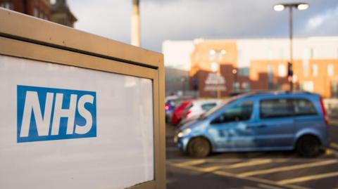 A car park with a sign next to it. The sign has the white and blue NHS logo on it.