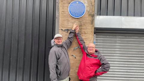 Two men in winter jackets point up to a blue plaque on a brick wall above them 