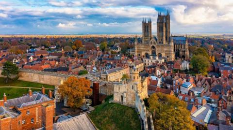 An aerial view across Lincoln. You can see the castle walls and the cathedral rising high behind them. The sky is cloudy and there are several houses and trees.