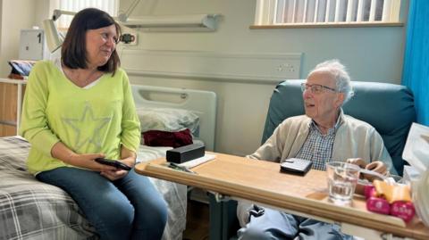 Roger sits in a padded chair next to a hospital style bed, which his daughter is perched on. A wooden table tray is in the foreground in front of his seat. They are chatting to each other. Roger is 92 and wears glasses, a checked shirt and beige cardigan. His daughter has a lime green sweater on with a silver star print and blue jeans.