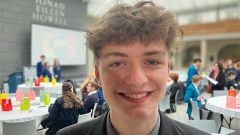 A teenage boy with curly brown hair smiling at the camera. He is wearing a grey school blazer and shirt. Behind him students are sat in groups in a workshop style on circular tables with bags on them. 