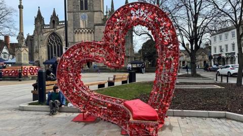 A large metal love heart structure adhorned with with red love hearts leans to the side and a curved seat protudes at the bottom. There is a red cushion to sit on. A church and war memorial can be seen in the background.