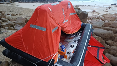 An inflated life raft on small granite boulders next to a sandy beach. It is about 8ft long, with an orange canopy, black rim and silver floor.