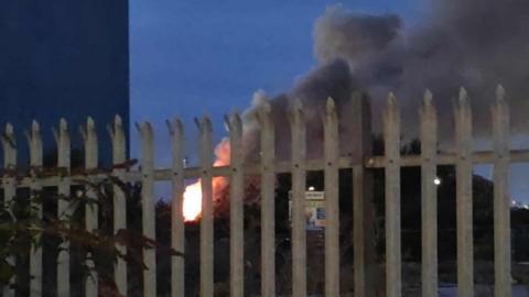 A fire burns inside a scrap metal yard in Gravesend, pictured through steel railings, with the dawn just breaking in the sky.