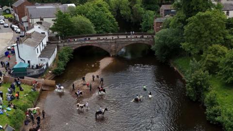An aerial image of the river in Appleby with several people bathing with their horses in the water. Spectators are on the grassy bank next to the river and others stand on a bridge looking on. In the distance, cars are parked on the road outside houses.