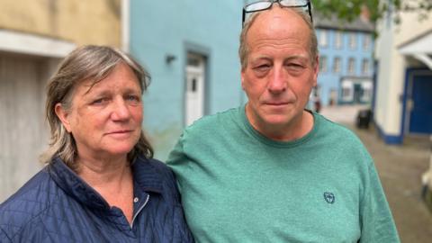 Woman and man stood outside on street looking at camera, with blurred background of town