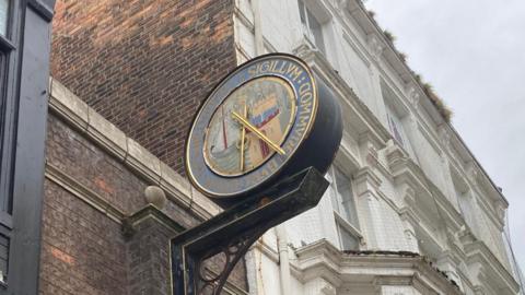 The exterior of a building with a round clock hanging from the side. The clock has two gold coloured hands and is attached to a decorative black metal bracket with gold detailing.The clock, which is wrong, reads 6:25.
