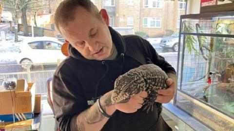 Dave Bigsby holding a large yellow snake and looking down at it, while stood inside his shop