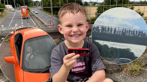 A young boy sits cross legged, smiling to camera while holding a red bus toy. A road can be seen in the background.