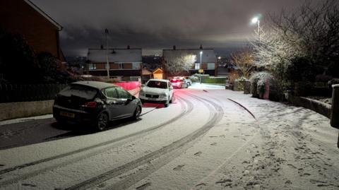 A street covered in snow. Two cars covered in snow are parked on the pavement. In the distance there are houses dusted with snow.