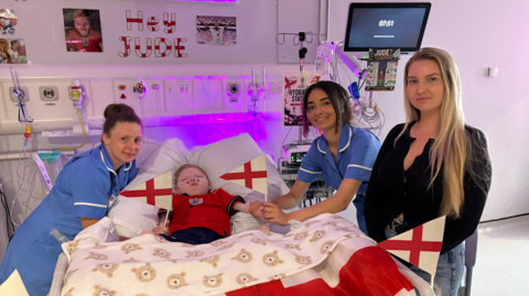 A young child lies in a hospital bed decorated with multiple England flags, including a large St George’s Cross flag hanging behind the bed and smaller flags attached to the bed rails. The child is wearing a red England football shirt and is covered by white bedding. Medical equipment, wires, IV stands, and wall-mounted hospital outlets surround the bed, indicating a hospital ward setting. The child has short, light-coloured hair and a nasal cannula providing oxygen. A chair and additional medical cables are visible in the background. The scene is lit with cool-toned hospital lighting.
