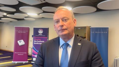 Deputy Lyndon Trott is wearing a blue tie and a black suit. He is standing in front of a Jersey Chamber of Commerce banner. 
