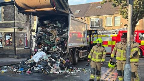 Refuse strewn on the road in the aftermath of a refuse wagon blaze with firefighters and a fire engine also in view