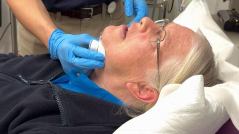 Natalie lies back on a hospital bed whilst the surgeon's gloved hand holds the white pad in place under her chin to test the implant.