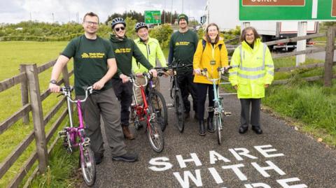 Five people, four men and one woman, with bikes posing for the camera on a cycle lane by a major road. Some are wearing dark green tops which say "Walk Wheel Cycle Trust". One person on the right, who does not have a cycle, is wearing a high-vis jacket. She has grey hair and glasses.