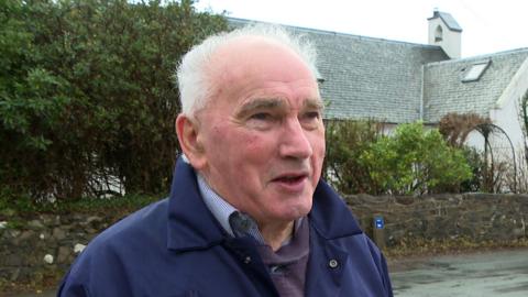 Teàrlach MacGilleBhraith na sheasamh aig taobh rathad agus balla is craobhan air a chùlaibh.