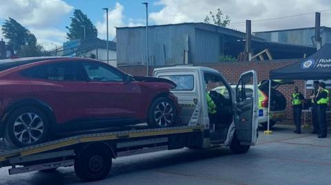 A red car is on the back of a white truck. There is  a building in front of the van and a police-branded black gazebo. There are three police officers wearing high visibility jackets standing underneath the gazebo and a marked police car alongside it with its boot open.