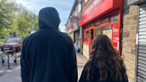 A boy in a black hoodie and a girl with curly brown hair look out a street, backs turned. To their right is a row of shops, to their left are trees and a car.