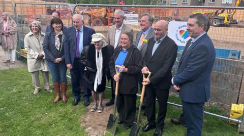 A group of representatives from Sir Bobby Robson's family, charity donors, clinical leads, Newcastle Hospitals (and the Newcastle Hospitals Charity), and the Sir Bobby Robson Foundation. Two are holding spades, and a patch of soil shows where the first ground has been broken on the project. Behind the group is a construction site surrounded by fencing where vehicles are working.