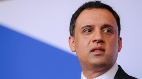 Anas Sawar stares into the distance. He has dark hair and is wearing a dark suit and white shirt. The background is blue and white.