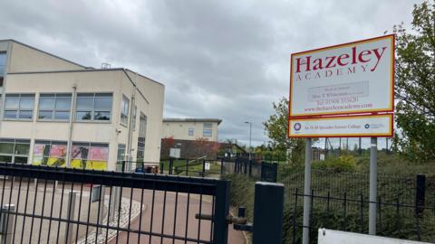 The school gates outside Hazeley Academy where there is a large white sign with the name of the school