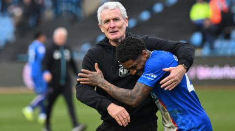 Carlisle manager Mark Hughes in his black tracksuit hugs his player Kadeem Harris at full-time after a crucial win