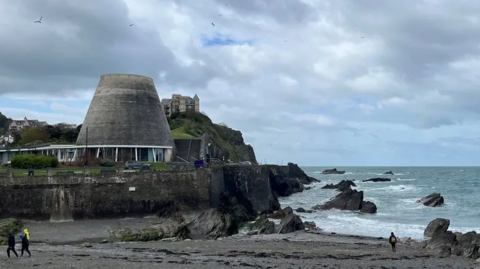 A view of the beach and the sea at Ilfracombe with rocks to the right and the conical tower of the Landmark Theatre on the left