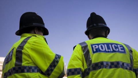 Two police officers are pictured from behind. They are both wearing high visibility jackets with the word 'police' written on the back. They are also both wearing black police hats.
