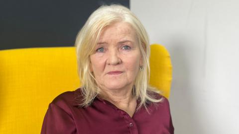 Catherine looking at the camera. She has shoulder length blonde hair and is wearing a burgundy satin blouse. She is sitting on a yellow chair.