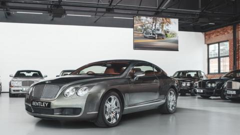 A Bentley showroom with a saloon car in the foreground and other cars lined up against the walls around it. A hanging screen with an image of an older car on a road is hanging from the ceiling behind it.