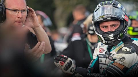Michael Dunlop, who is in his leathers and has his helmet on. The visor of the helmet is up. Richard 'Milky' Quayle is standing to his left beckoning someone else over.
