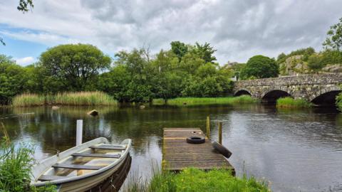 Clouds and a little blue sky over a rural river, with a jetty and boat in the foreground. 