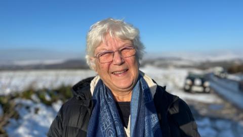 An elderly lady with short-cropped hair squints with the sun in her eyes.