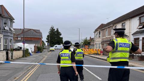 Three police officers stood by police tape inside a police cordon. They are wearing high vis jackets and black police hats. You can see houses in the background.