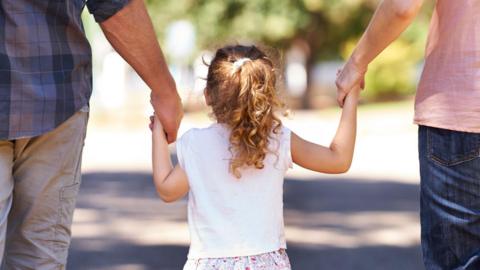 Young girl standing in the middle of her mother and father, holding both of their hands. They're all facing away from the camera.