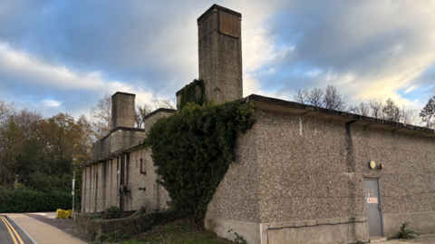 A 1950s building covered in pebbledash and a climbing green plant up a portion of one wall