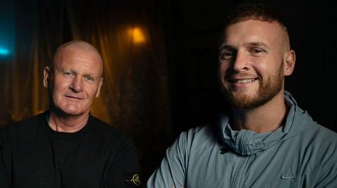 Andrew and Macaulay Dodd pictured standing next to each other. Both looks at the camera and smiles. Andrew wears a black top and Macaulay a light blue sports zip-up top. It is a head and shoulders shot of them.