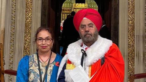 A bearded man wearing a red robe and red turban, and a woman wearing glasses and a blue dress, standing in front of an opulent stone door with gold trimming.