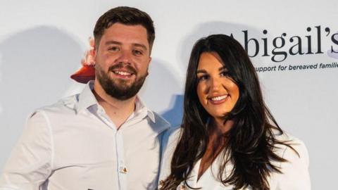 Matt Knight on the left has dark short hair and a short beard, he is wearing a white shirt. Hannah Spearing next to his on the right has long dark hair and is wearing a white blazer. They're both smiling.