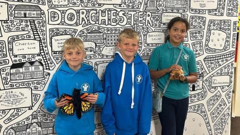 Two young boys and a young girl standing against a black and white decorated wall. One of the boys holds a cuddly toy butterfly and the girl holds a cuddly toy tortoise