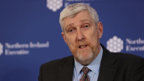 John O'Dowd is an older man with short, white hair. He is speaking at a press conference in front of a blue 'Northern Ireland Executive' wall display. He is wearing a black suit jacket, a blue shirt and a patterned tie.